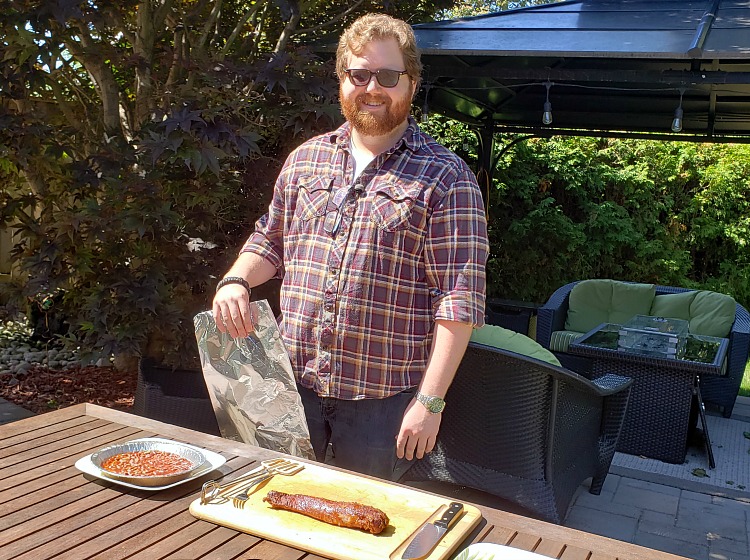 A man stands outside with a barbecued pork tenderloin on a cutting board in front of him.