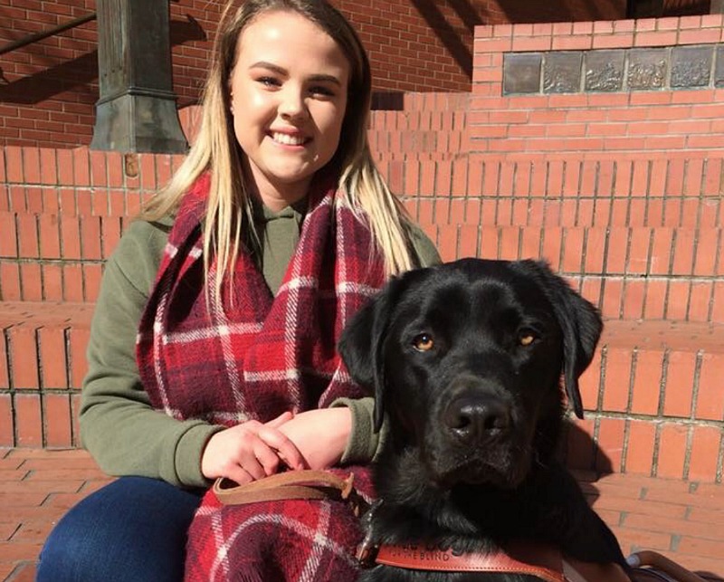 A woman smiles into the camera. A guide dog sits next to her.