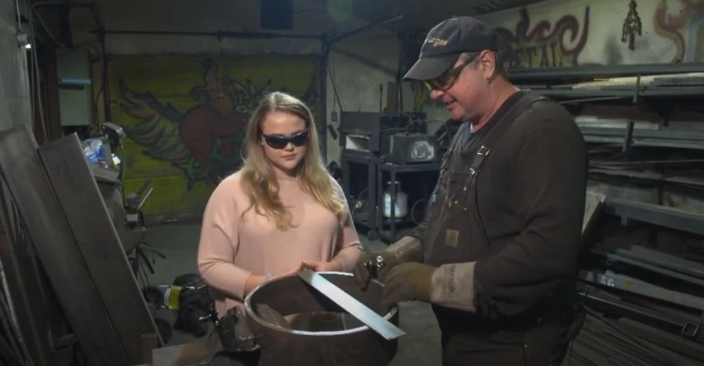 A woman and man stand at an anvil, shaping metal.