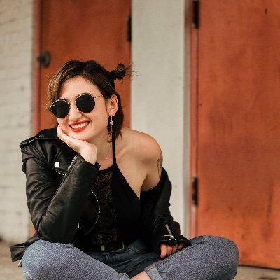 Jess Rauchberg sits cross-legged wearing a leather jacket and sunglasses. 