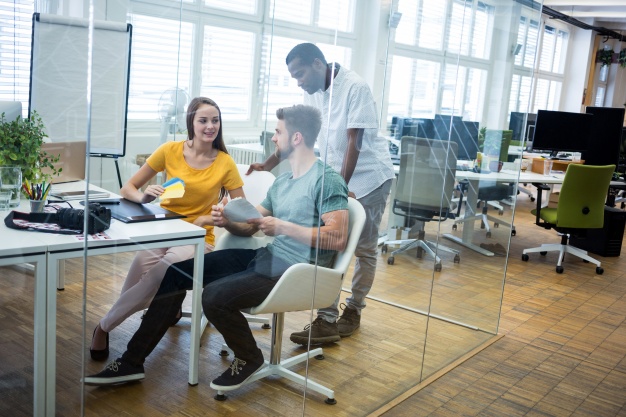 Image shows office a woman and a man talking to each other while seated at a table. A second man is behind them, leaning on both of their chairs. They are in an enclosed glass meeting room within a larger office.  