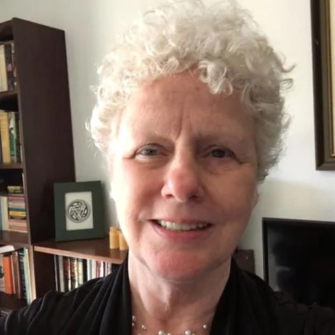 A woman with grey curly hair looks ahead, with bookcases in the background