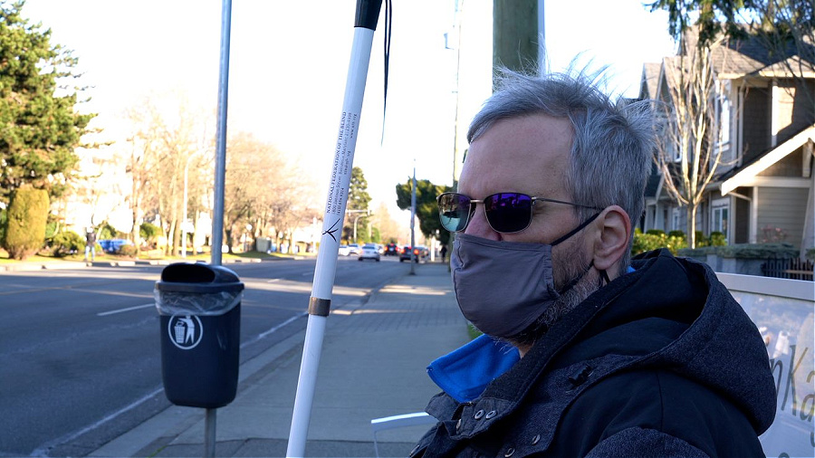 A man, wearing a surgical mask, stands at a bus stop. He is holding a white cane.