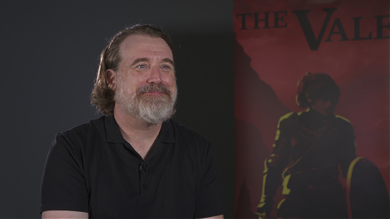 A man sits in front of a poster for The Vale