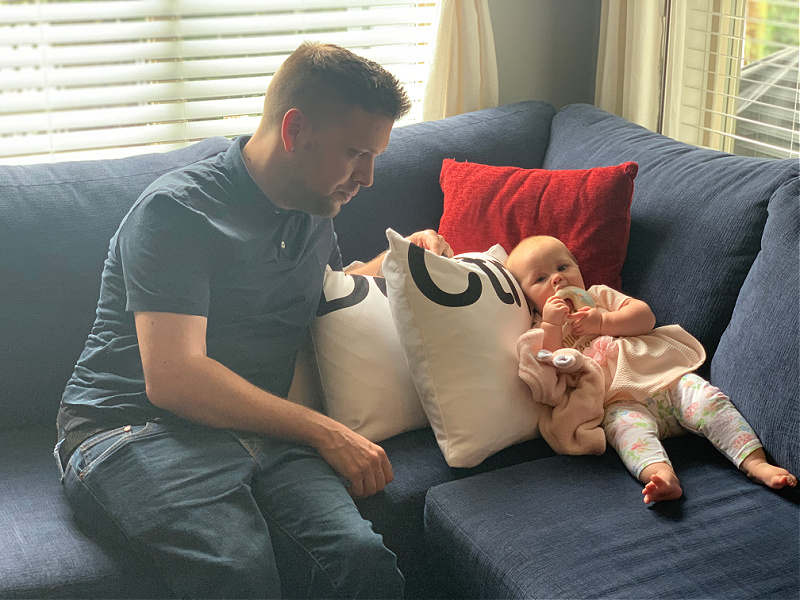 Marco Pasqua, seated on a couch, looks down at his infant daughter.