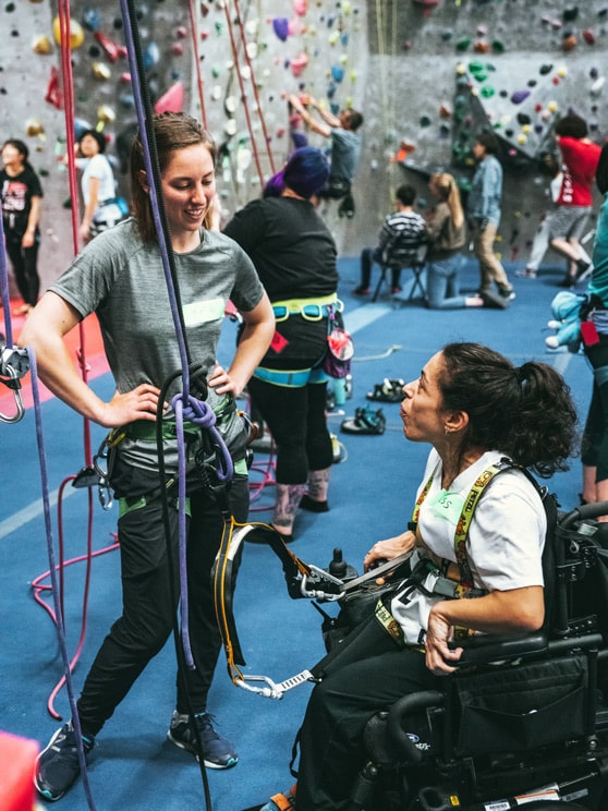 A rock climbing instructor helps a woman using a wheelchair get rigged up to climb a rock wall 