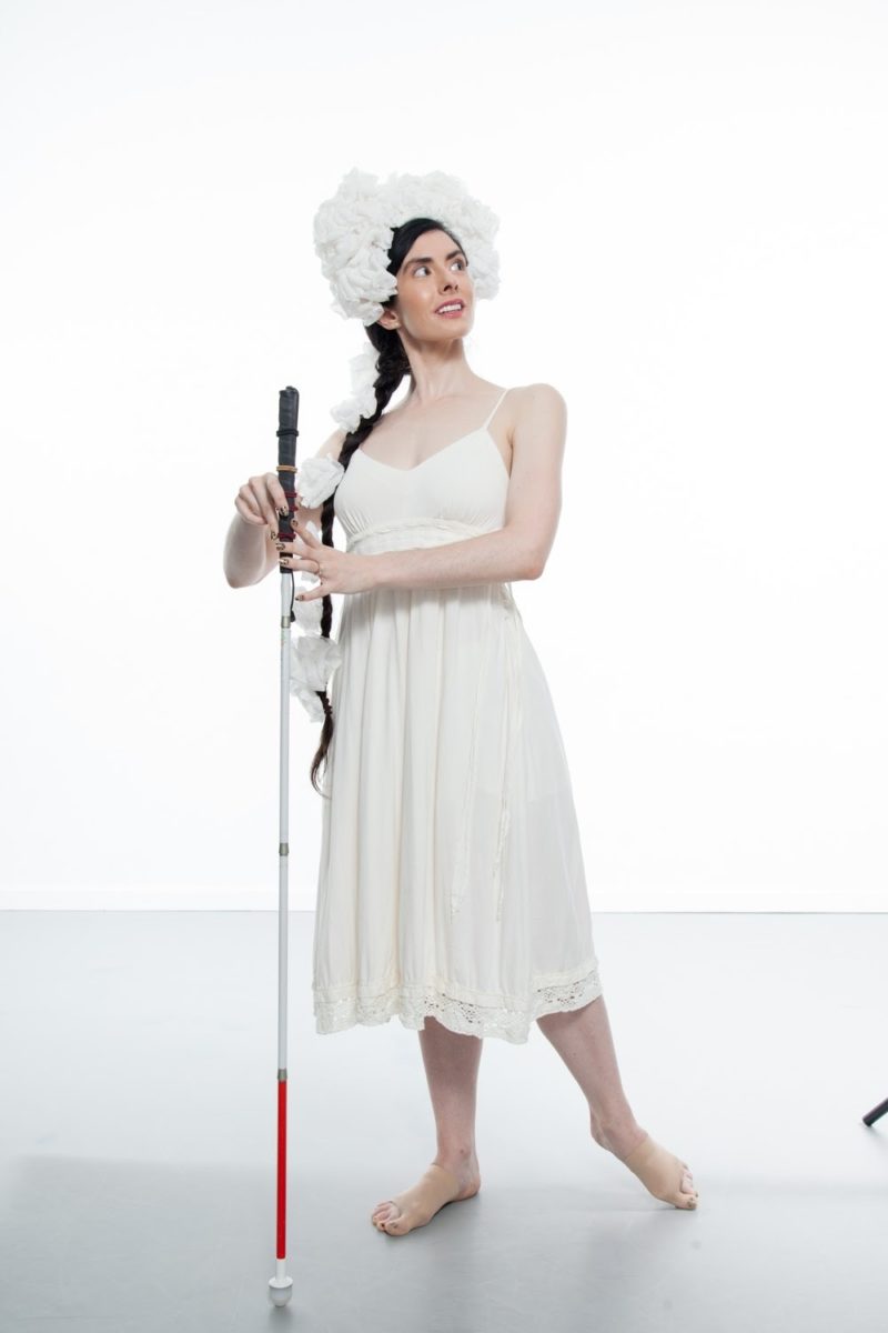 A woman in a white dress and matching floral bonnet poses with her cane on stage.