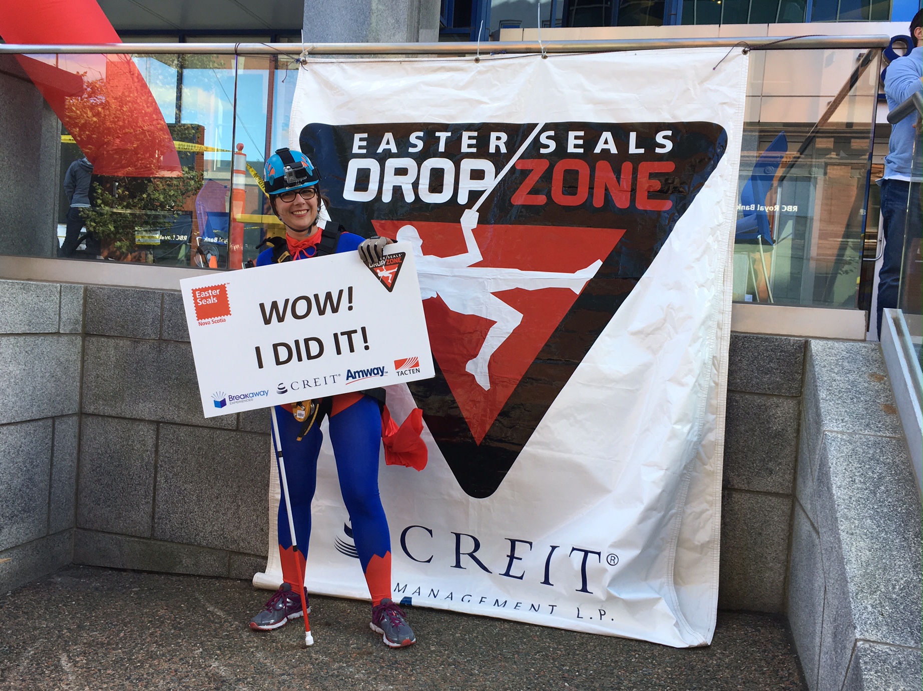 Laura Bain hold up a sign that says "Wow! I did it!" as she stands in front of a banner that reads Easter Seals Drop Zone. She's holding her white cane and wearing a blue helmet. 