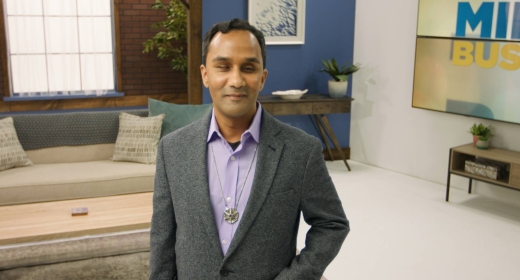 The image shows a man in a gray blazer standing in a well-lit, modern room. The text in the foreground reads “MIND YOUR OWN BUSINESS,” with “MIND” in bold blue letters, “YOUR OWN” in smaller white text, and “BUSINESS” in bold yellow letters. The setting appears professional, and the man is smiling slightly, giving a calm and confident impression.